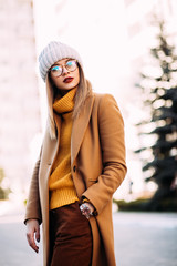 City lifestyle.Outdoor portrait of young beautiful woman Model wearing stylish glasses,mustard sweater, beige coat and grey knitted hat