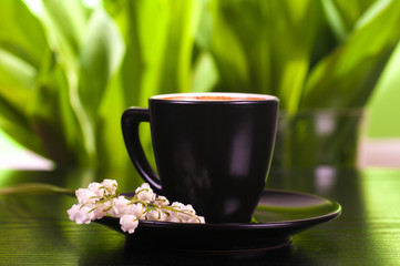 coffee in a black cup with a saucer, lilies of the valley on a saucer, green leaves background