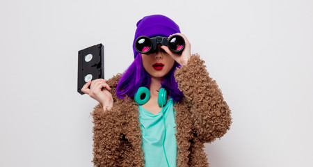 Beautiful young girl with purple hair in jacket holding VHS cassette and binocular on white background.