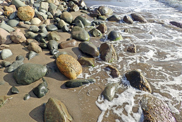 Various shapes and sizes of rocks, stones and pebbles in colors of coral, jade, rust, and tan in bright sunlight scattered on a dark brown sandy beach washed with the foam of the sea surf.