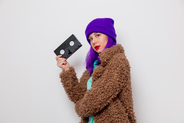Beautiful young girl with purple hair in jacket holding VHS cassette on white background.