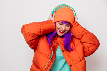 Beautiful young girl with purple hair and in orange jacket listen music in headphones on white background.