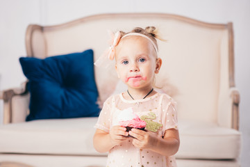 children's birthday. funny two-year-old Caucasian girl in pink dress standing to bedroom of house the background of couch and eating, holding cake dessert. Face and mouth smeared, smeared with cream