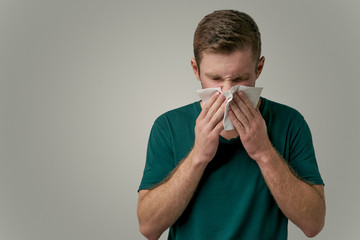 An unhealthy man feels bad. The guy in the green t-shirt blows his nose and sneezes into the handkerchief. Isolated on a gray background in the Studio