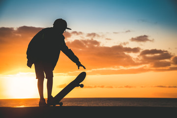 Silhouette of young boy plays with skateboard at sunset near Backlight of a skater with hat standing in a orange sky with golden reflections on the ocean. Teenager playing with a foot on the board