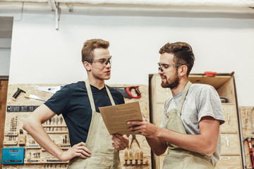 Two young men in aprons and protection goggles smiling and discussing documents while standing in...