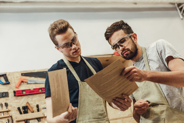 Two attractive guys in goggles looking through documents while standing in modern joinery together