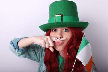 Red hair girl wearing a green Leprechaun's hat making a face with a fake moustache and holding a flag of Ireland