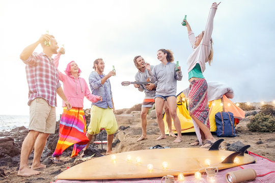 Happy friends having fun playing guitar and dancing on the beach - Group of young people making party drinking beers while camping with tent - Travel, vacation and youth holidays lifestyle concept