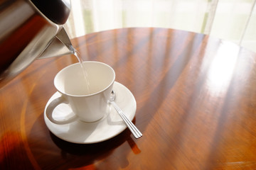 Filling hot water from kettle to white empty glass with saucer and metal spoon on wooden glossy round table nearby the curtain at window with sunlight