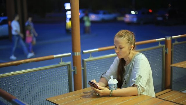 young single woman sitting in the evening in a summer cafe on the street , listening to music on smartphone and have dessert.