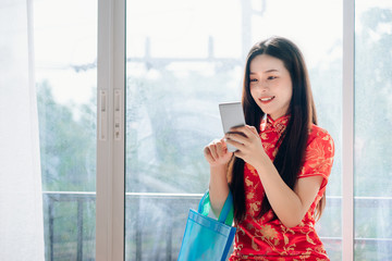 Smile of Portrait Beauty Asian Woman with Chinese dress,Qipao,Cheongsam Chinese New Year Using Smartphone Technology Shopping Online and hold Bag Sitting near Window in Clean White Room,Copy Space.