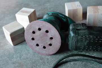 Dusty electrical orbital sander with cable, sanding disc and natural raw wooden cubes on grey grunge table. Carpentry equipment and making wooden toys diy concept.