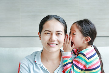 Happy Asian child girl sharing a secret to her mom in the living room at home. Kid girl whispering gossip a something to mother ear.