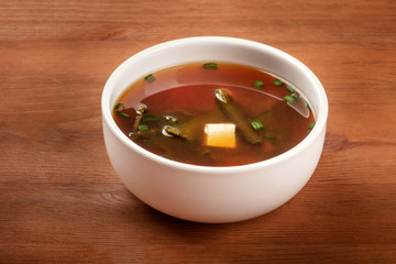 A photo of a bowl of miso shiru soup with tofu, scallions, and wakame seaweed, on a dark rustic wooden background with a place for text