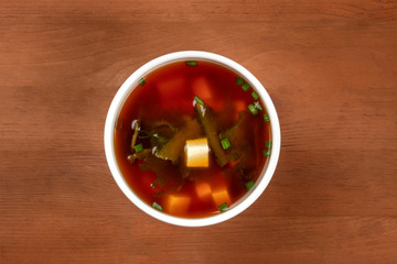A photo of a bowl of miso shiru soup with tofu, scallions, and wakame seaweed, shot from above on a rustic wooden background with copy space