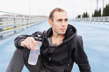 Obraz premium Close-up of a young man personal trainer in a gray suit drinking water from his bottle after a fat burning morning street workout. Concept of water balance replenishment during workouts