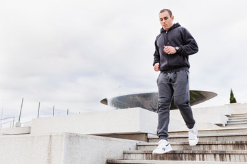 Young slim man a runner in a gray tracksuit and sneakers moves up the steps around the sports stadium in early cloudy summer morning. Concept of energy and aerobic exercise. Copyspace