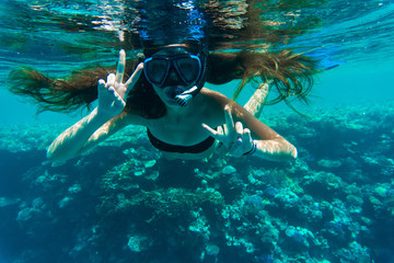 Young woman snorkeling making peace signs underwater near coral reef