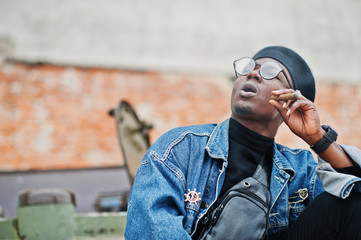 African american man in jeans jacket, beret and eyeglasses, smoking cigar and posed against btr military armored vehicle.