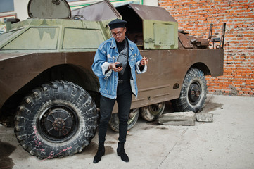 African american man in jeans jacket, beret and eyeglasses, smoking cigar and posed against btr military armored vehicle, with mobile phone at hand.