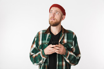 Portrait of bearded guy wearing hat and plaid shirt using mobile phone, while standing isolated over white background