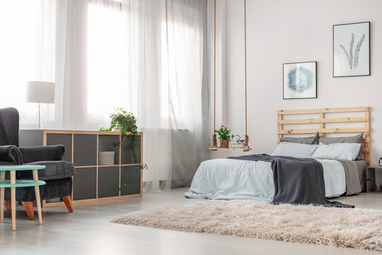 Fluffy white carpet on the floor of trendy bedroom with king size bed with wooden headboard, posters on the wall and fashionable swing as nightstand