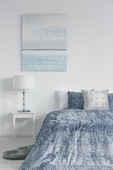 Vertical view of white stylish lamp on bedside table in luxury bedroom interior with velvet blue bedding and painting on the wall