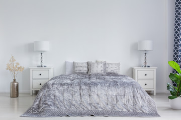 Grey bedding on king size bed in stylish bright bedroom interior with white bedside tables and lamps, real photo with copy space on empty white wall - obrazy, fototapety, plakaty