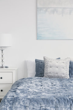 Closeup of white stylish lamp on bedside table in luxury bedroom interior with velvet blue bedding and painting on the wall