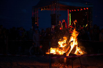 Large burning bonfire with soft glowing flame and sparkles flying all around. Romantic summer evening, people relaxing and enjoying calmness at the festival on seaside. Stage, singers in background