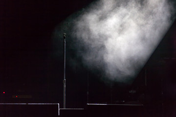 Microphone holder standing on a black dark scene, smoke illuminated by white spotlight at the side, below are stage monitors. Microphone stand on stage.