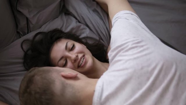 Sensual man and woman lying in the bed with grey sheets. Intimate young couple enjoying foreplay on bed. Top view