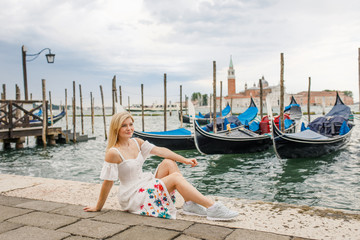 Fototapeta na wymiar Young beautiful girl in white dress sits near the water overlooking the gondolas and beautiful Venice