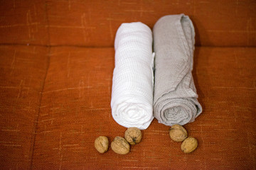 Obraz na płótnie Canvas Macro shot of rolled, folded snow white and grey natural linen sauna and bath towels next to small walnuts on orange background, on massage table. Beautiful home decor, gift for newlyweds for wedding 