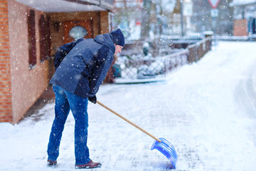 Man with snow shovel cleans sidewalks in winter during snowfall. Winter time in Europe. Young man in warm winter clothes. Snow and weather chaos in Germany. Snowstorm and heavy snowing. Schneechaos