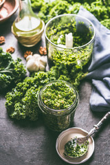 Raw kale eating recipes. Green kale pesto in glass on dark rustic kitchen table background with ingredients, top view. Kale preparation. Healthy detox vegetables . Clean eating and dieting concept. - obrazy, fototapety, plakaty