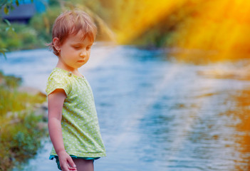 Naklejka premium Adorable little child girl playing and having fun by leaves near mountain river on warm and sunny summer day. Holiday, rest, happy childhood, games, nature concept