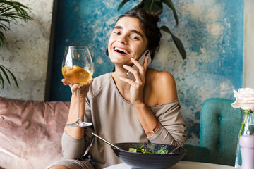 Beautiful young woman sitting in cafe indoors drinking white wine eat salad talking by mobile phone.