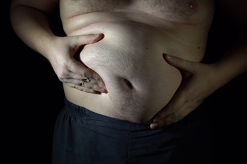 Unhealthy man checking his big belly in front of dark background.