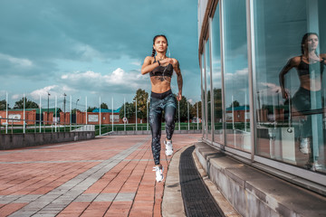 Fototapeta na wymiar Girl athlete in jump, runs for morning jog. In summer city. In hands of smartphone and headphones. Tanned skin and tattoos. View from front on background of glass windows. Free space for text.