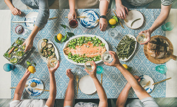 Mediterranean style dinner. Flat-lay of table with cooked salmon, starters, bread over blue table cloth with hands holding drinks, sharing food, top view. Holiday party concept
