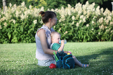 A young mother wearing a dress is sitting on the lawn in the garden with her baby on a sunny summer day