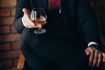 A man in a black suit sits on a chair and holds a glass of cognac. Stylish bride's morning. 