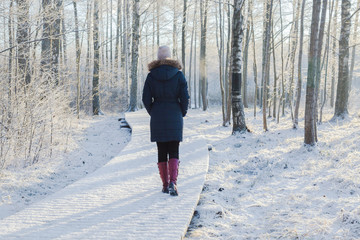 Young adult woman alone slowly walking on snow covered boardwalk after snowfall. Enjoying fresh air in park. Peaceful atmosphere in winter day. Back view.