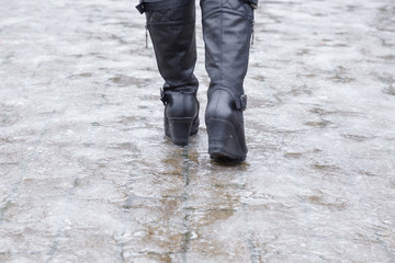 Young woman's legs in black leather boots walking on sidewalk in wet, warm winter day. Pavement...