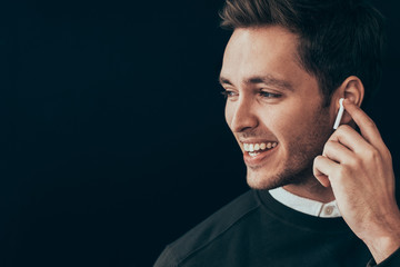Closeup horizontal portrait of young handsome man smiling and have a call with a colleague isolated on black background holding wireless earphone with finger. Businessman using wireless headphone. - obrazy, fototapety, plakaty
