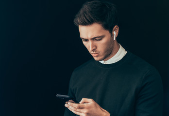 Portrait of young handsome man texting messages from his smart phone against studio black background, with wireless earphones. Caucasian businessman using wireless headphones