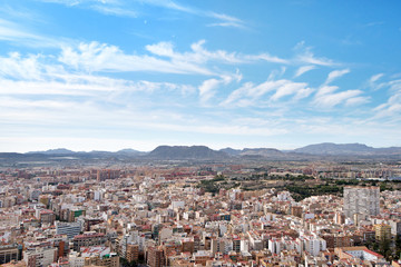 Fototapeta na wymiar Panoramic view of city from Santa Barbara Castle in Alicante, Spain. Block apartment buildings, parks, roads, houses, palm trees. Beautiful mountain landscape in background, blue sky 