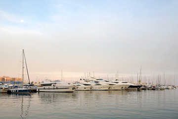 Fototapeta na wymiar Beautiful port of Alicante, Spain at Mediterranean sea. Luxury yachts, ships, ferries and fishing boats sailing and standing in rows in harbor. Rich people traveling around the world. Sunset evening 